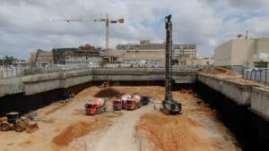 פועלי בניין עובדים באתר בנייה. 3 משאיות בטון, שופל ומכונת קידוח. ברקע רועים עגורן בעבודה